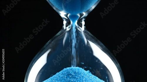 Close-up of a glass hourglass with blue sand flowing, symbolizing the passage of time against a black background
