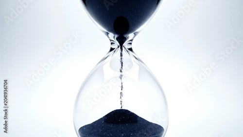 Time passing quickly as black sand flows through a clear glass hourglass on a bright background