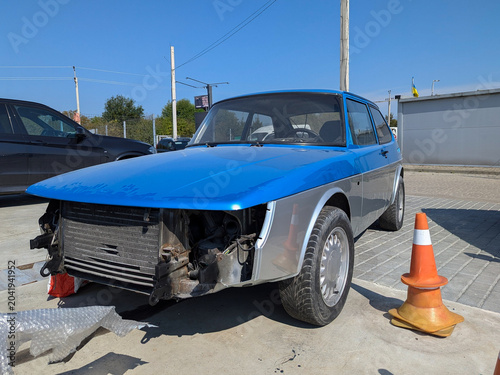 A heavily damaged blue vintage car with its hood open and front end destroyed, standing next to an orange traffic cone in an outdoor lot.