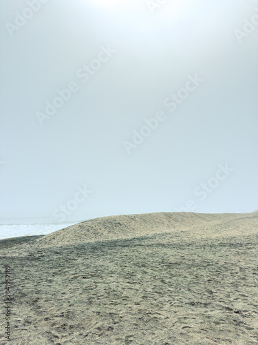 Tablou pe pânză Waves reach shore while sun shines briefly over beach during midday with light f