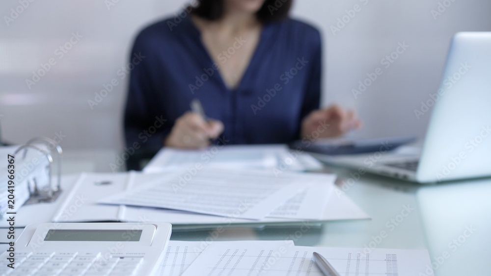 Fototapeta premium Magnifying glass, white calculator, pen and documents are lying on accountant's desk. Businesswoman wearing blue dress is working with documents on the background. Business and audit concept