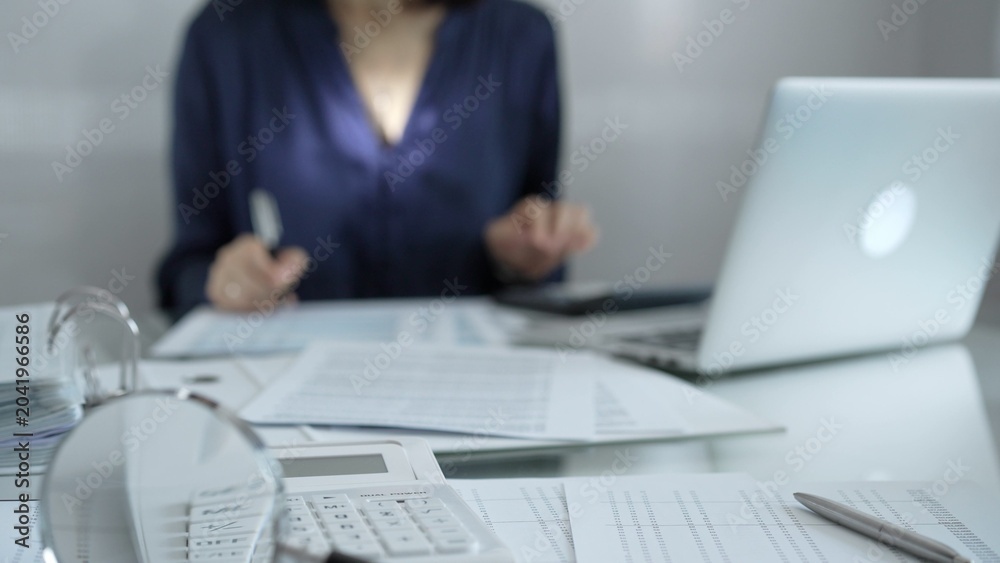 Fototapeta premium Magnifying glass, white calculator, pen and documents are lying on accountant's desk. Businesswoman wearing blue dress is working with documents on the background. Business and audit concept