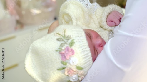 A soft close-up shows a newborn in a cream knitted bonnet resting against a mother in white, filmed with shallow focus and gentle motion. It suits bonding, newborn care, warmth, and home lifestyle.
