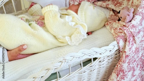A gentle close-up shows a mother laying her newborn into a white wicker cradle, supporting the body with slow careful motion. It suits baby care, bedtime routines, bonding, and tender home life.