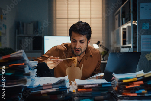 Foto Young Latin businessman eating takeout instant noodles and reviewing document on laptop at desk in dark night office