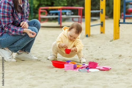 Toddler playing with toy dishes in sandbox while mother watches at playground