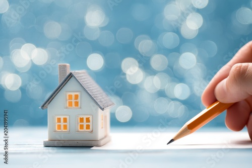 Miniature house on blueprints with a hand holding pencil. Building model close-up with contract paper, a new home conceptual. Plans and project documents. Architectural layout. Real estate background