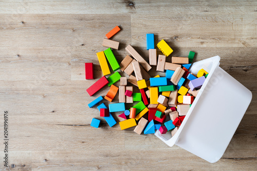 top view of colorful wooden toy blocks spilled from a white plastic storage box onto a light wooden floor