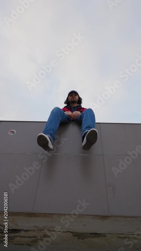 Person sitting on edge of urban structure viewed from low angle against cloudy sky. Concept of contemplation, solitude and urban lifestyle. Vertical video