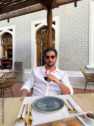 Handsome dark-haired man in a linen shirt sitting at a local Greek island restaurant, holding a glass of wine, elegant pose, sunset ambiance, seaside dining, Mediterranean lifestyle