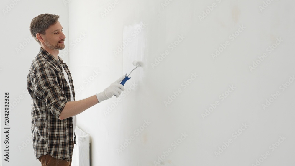 Fototapeta premium Middle aged man wearing checked shirt and protective gloves, is painting a white wall with a paint roller, working on a home renovation project, creating a fresh and clean look