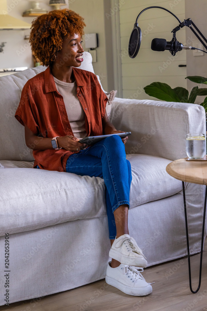 Obraz premium African American woman sitting on sofa wearing rust shirt holding tablet speaking into mic at home