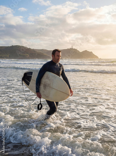 A middle-aged surfer walks through shallow water before entering the sea to surf.
