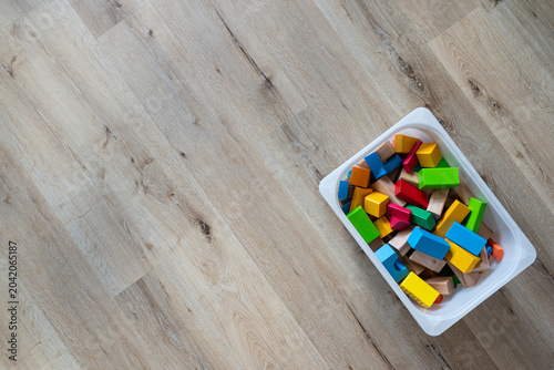 top view of colorful wooden toy blocks organized inside a white plastic storage box on a light wooden floor, featuring copy space