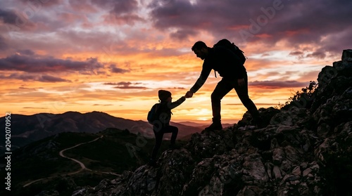 Father and son silhouettes holding hands while climbing a rocky mountain peak during a vibrant orange sunset in the evening