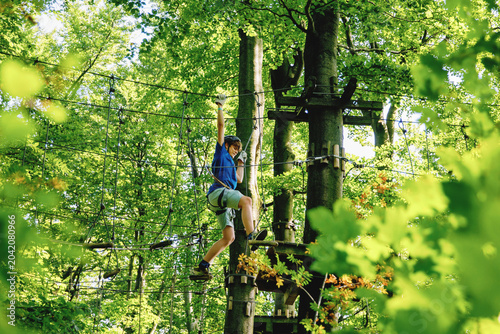 Teenage boy climbing rope course in forest adventure park, child wearing helmet and harness enjoying outdoor challenge, real life.