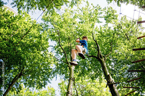 Teenage boy climbing rope course in forest adventure park, child wearing helmet and harness enjoying outdoor challenge, real life.