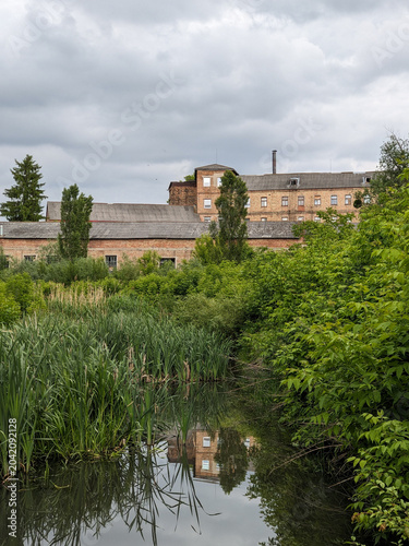 Building reflected in calm water surrounded by lush vegetation beneath an overcast sky.