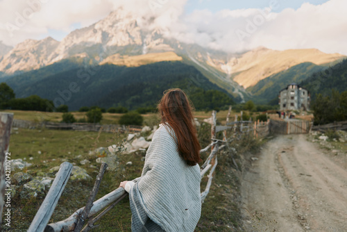 Obraz na płótnie A young woman in a light shawl stands by a wooden fence, gazing toward distant mountains in a quiet rural landscape