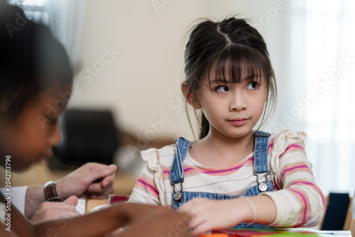 Tableau sur toile Asian elementary girl listens beside diverse classmate while teacher guides bloc