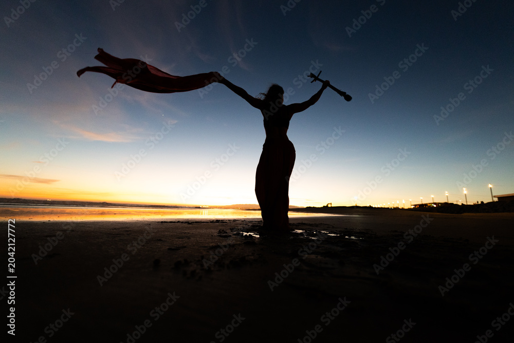 Fototapeta premium Silhouette of a woman in a flowing dress holding a stick against a colorful sunset at the beach, with reflections on the wet sand and distant lights