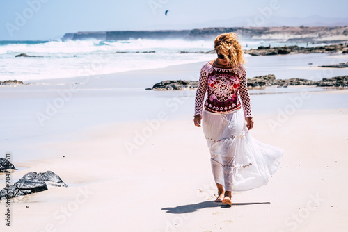 Quadro em tela Woman walking alone on a sunny sandy beach near rocky shore, wearing white skirt