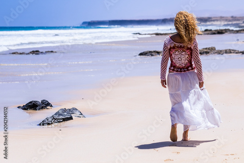 Woman walking on a sandy beach in a white flowing skirt and crochet top, strolling along the ocean shoreline for a peaceful sunny seaside summer escape