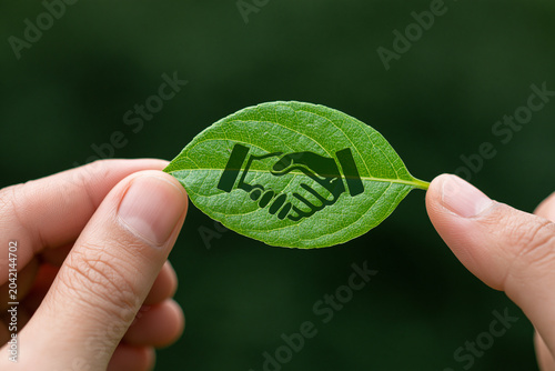 Close-up of human hands holding a fresh green leaf with a handshake symbol cutout representing environmental partnership, sustainable business deals, and corporate social responsibility.