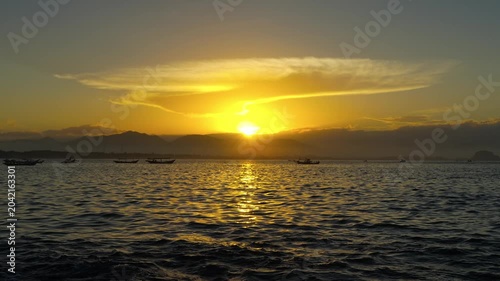 Dramatic sunset sky filled with golden light illuminating the ocean surface and traditional fishing boats near the mountain bay. Papuma Beach, East Java.