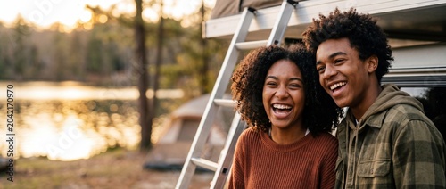 Black couple camping lake sunset laughing together during summer road trip adventure with rooftop tent vehicle; happy African American young man and woman enjoying outdoor adventure and freedom.