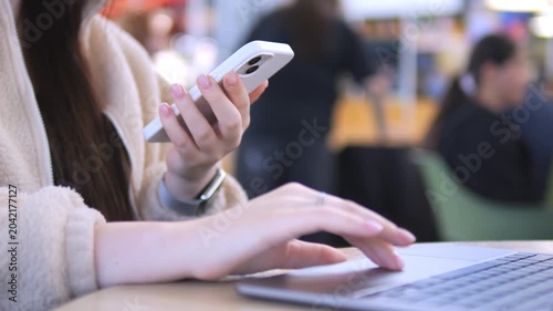 Young woman focused on smartphone in busy cafe. Hands scroll, fingers tap, eyes concentrate. Laptop nearby suggests multitasking. Perfect for tech, social media, or modern work themes