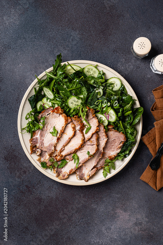 Slices of pork meat roast served with green vegetables salad in plate, top view