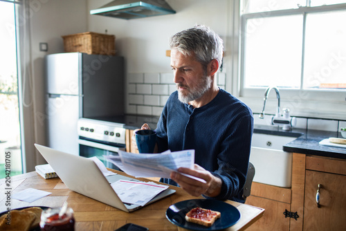 Fotografie Mature man reviewing household bills with laptop in home kitchen