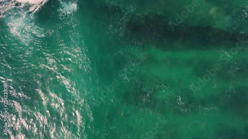 Aerial shot of the ocean with blue colours, waves doing  patters, rocks, sand and beach. Beautiful scenery