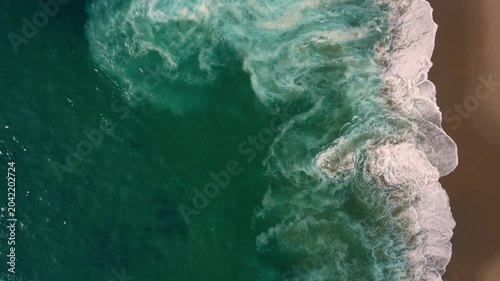 Aerial shot of the ocean with blue colours, waves doing  patters, rocks, sand and beach. Beautiful scenery