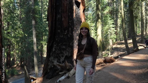 Smiling young woman wearing a yellow beanie walking on a dirt path in a sunny forest, admiring the giant ancient trees of sequoia national park in california during her vacation