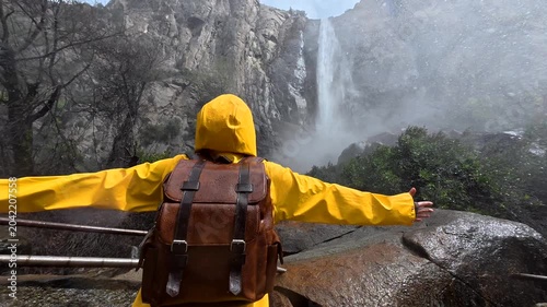 Happy female tourist wearing a bright yellow raincoat and backpack opens her arms wide, feeling the mist from the powerful bridalveil falls waterfall in yosemite national park, california