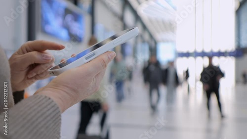 Hand holds tablet in busy transit hub. Person focused on screen, ignoring background movement. Blurred travelers walk past in hallway. Ideal for travel, technology, or modern commuting themes