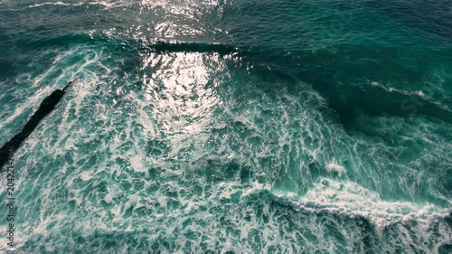 Aerial shot of the ocean with blue colours, waves doing  patters, rocks, sand and beach. Beautiful scenery