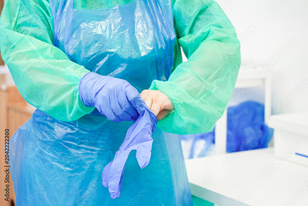 Fototapeta premium A medical professional wearing a protective gown puts on blue nitrile gloves in a clinical setting to ensure hygiene, sterility, and safety protocols during daily healthcare work
