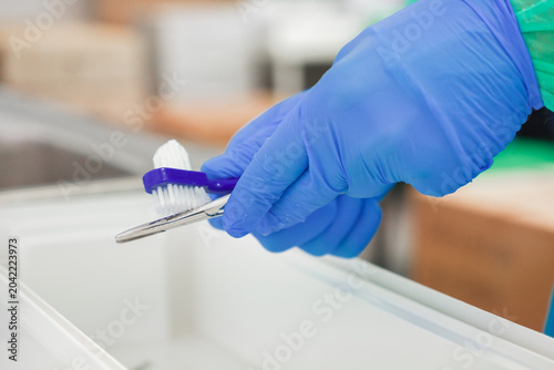 Gloved hand cleans a surgical instrument with a small scrubbing brush in a sterile hospital setting, representing the essential maintenance of medical equipment for patient safety