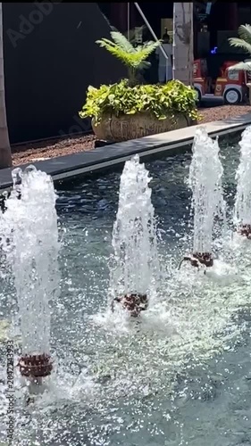Water fountain with vertical jets creating splashes and ripples with lush planter and seating area in the background evoke relaxation, summer, and public space design,4K.