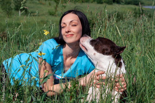 Happy woman closing eyes while her dog licks her face in the green grass outdoors. pet affection, summer field walk