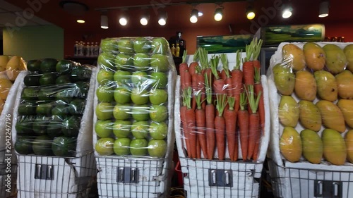 Variety of fresh produce, including carrots, mangoes, avocados, and green apples, wrapped in plastic and displayed in baskets at a local grocery store or market for sale