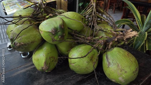 Large bunch of fresh young green coconuts resting on a dark wooden cart at an outdoor market, ready for sale. The tropical fruit is a popular source of refreshing and nutritious water