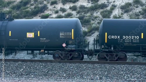 Two black tanker train cars move on railroad tracks with gravel below and sandy hills with green shrubs in the background on an overcast day in a remote outdoor setting