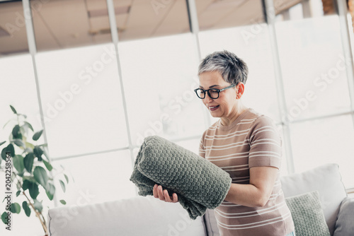 Photography Senior woman folding a soft green blanket at home in a bright, cozy living room,