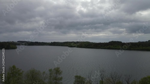  Aerial drone footage capturing the serene landscape of Stokka lake in Stavanger, Norway. The scene features a vast, calm body of water surrounded by lush greenery, captured at midday.