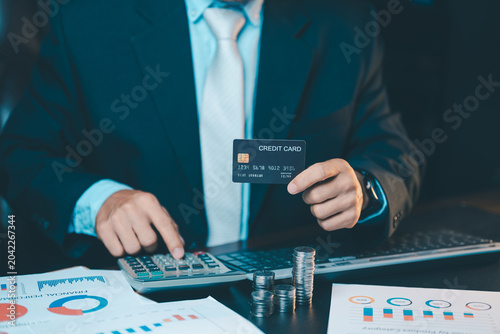Close up of businessman hand holding a black credit card and using calculator with coins stack on table. Financial planning concept.