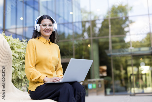Young woman smiling, wearing headphones and clear glasses, working or studying on a laptop while sitting on a modern bench outdoors in front of a glass building